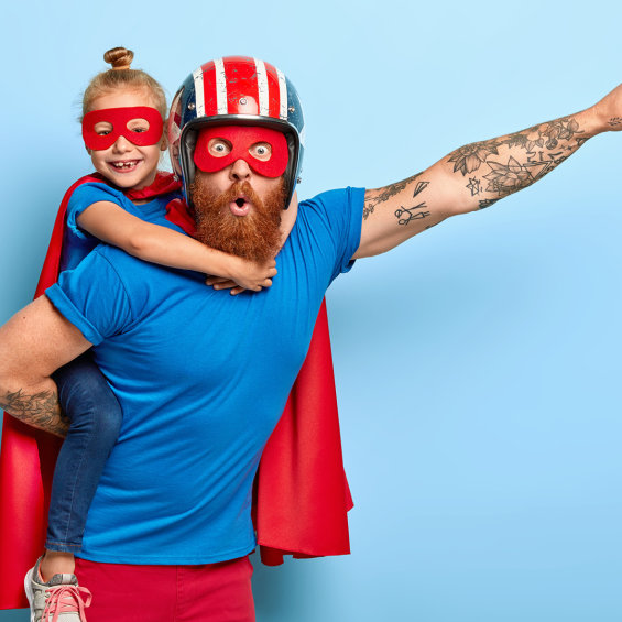 Tochter auf dem Rücken ihres Vaters, beide im Superhelden-Look mit Maske und Cape.
